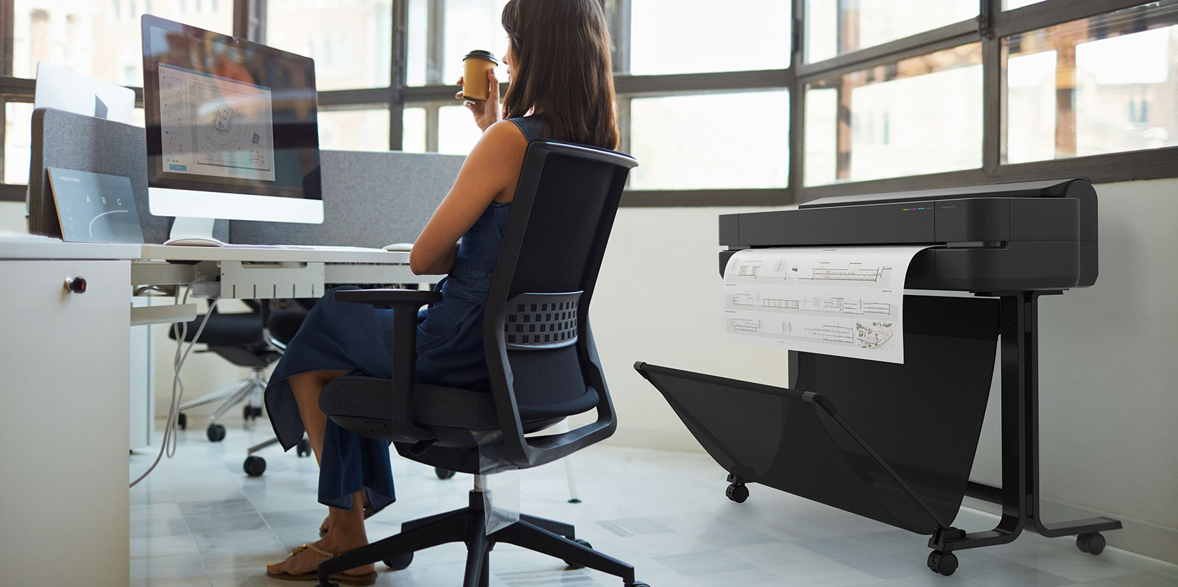 Person working with an HP computer on a desk in an office next to an HP DesignJet plotter