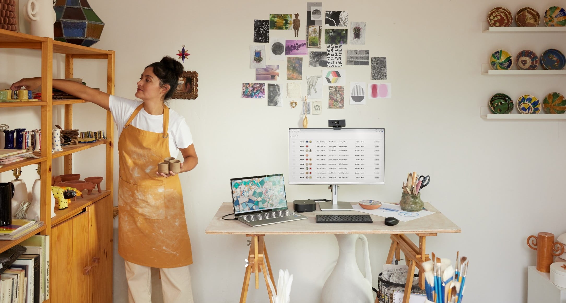 A woman storing some pottery cups aside her HP laptop connected to an HP monitor with an HP webcam and wireless keyboard and mouse.