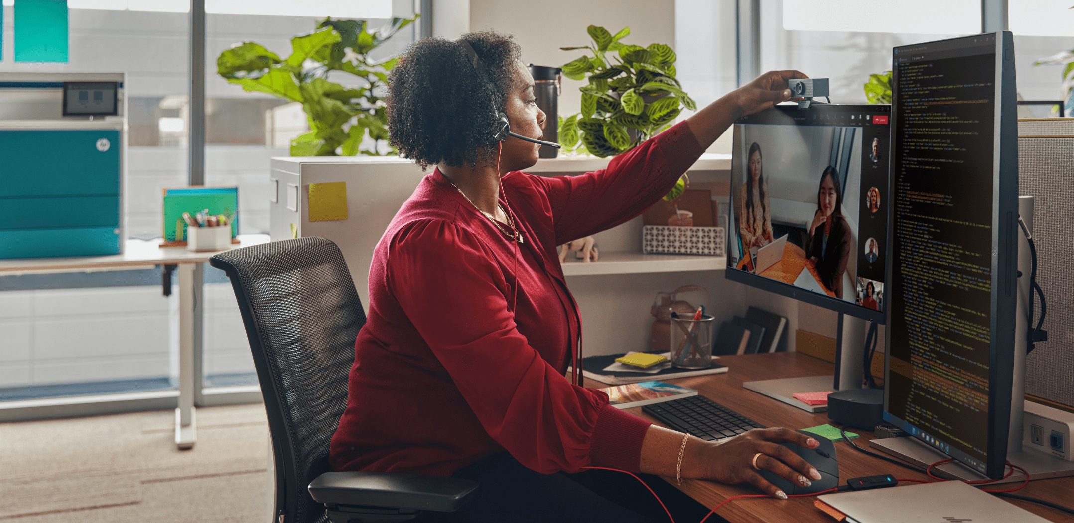 Woman seated at desk in open plan office with Poly Blackwire 3300 series headset hanging over her monitor