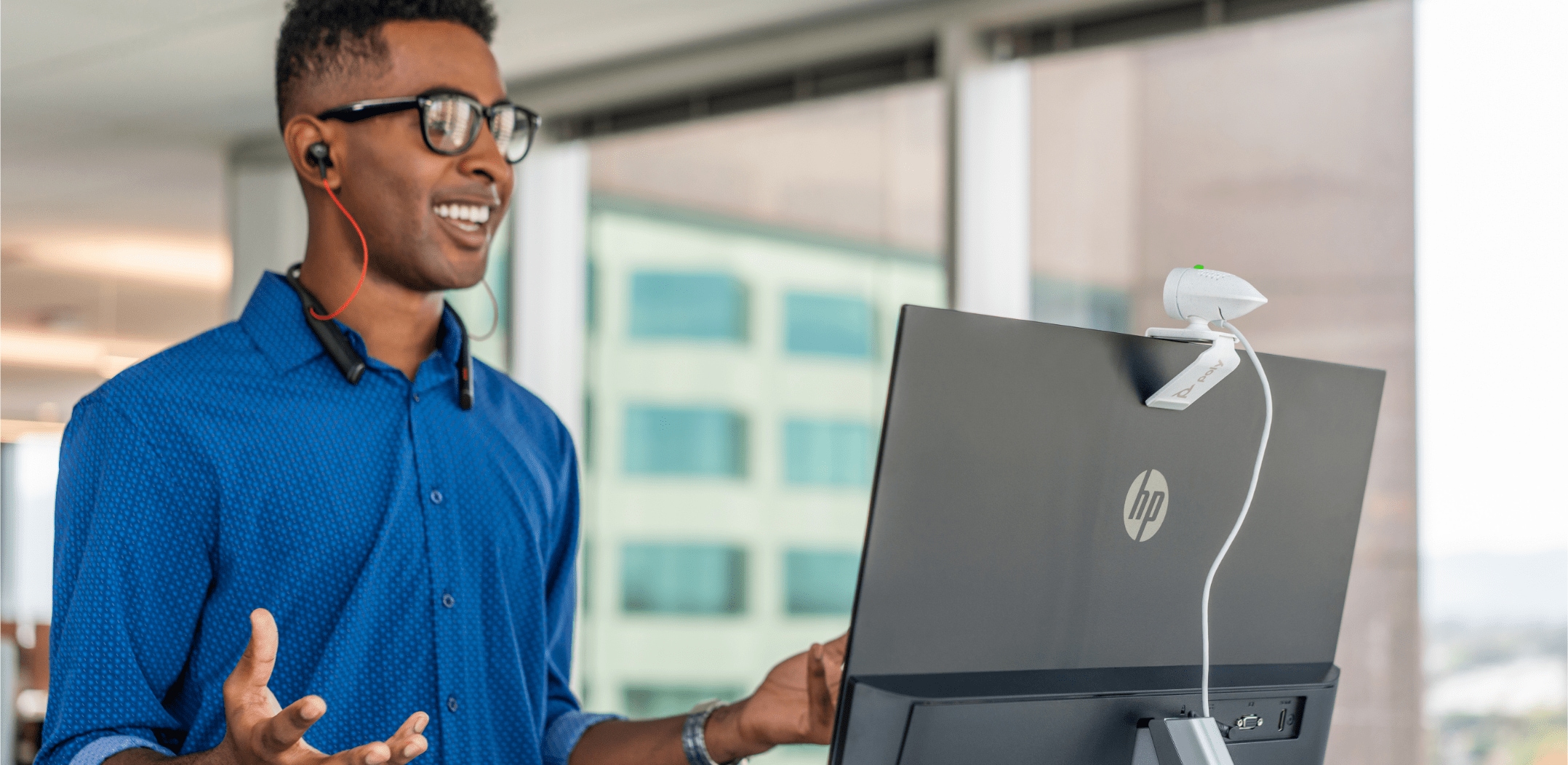 Man at standing desk in open plan office, engaged in a video call using Poly Studio P5 USB webcam with Voyager 6200 UC headset