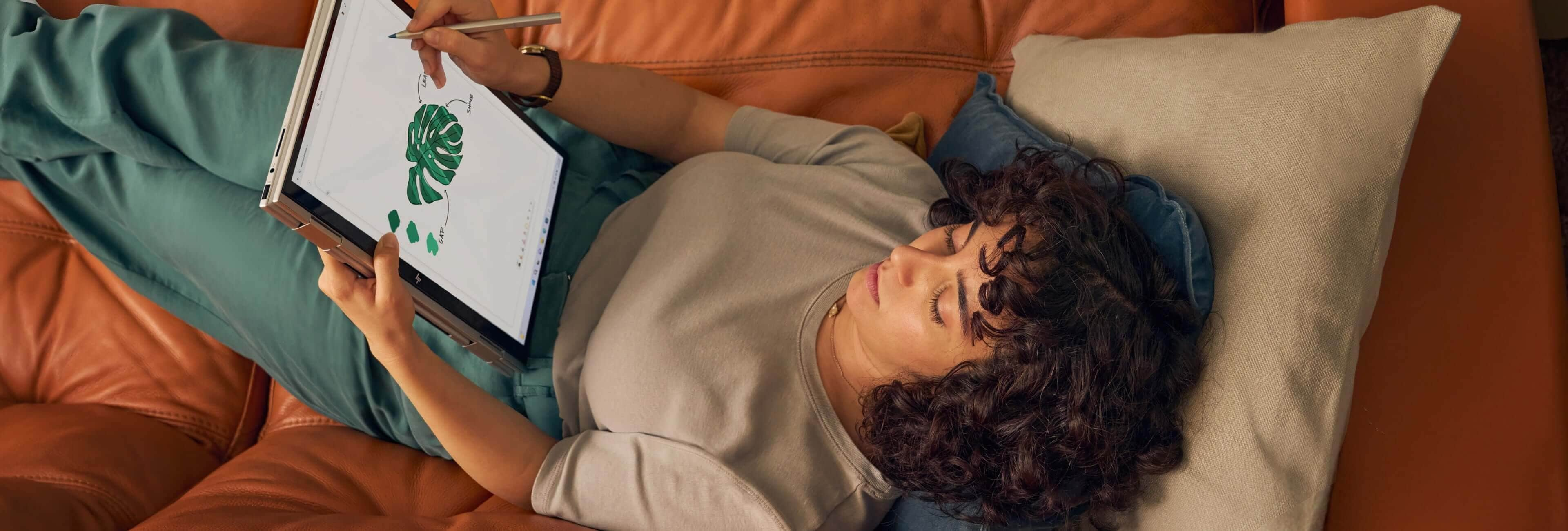 A woman lying down on her couch while drawing on her HP Envy laptop in tablet mode.