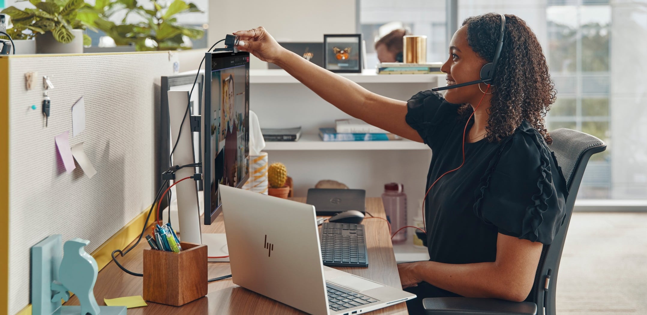 Woman seated at her office desk, wearing Poly Blackwire 3200 series headset while adjusting webcam on monitor during a video call