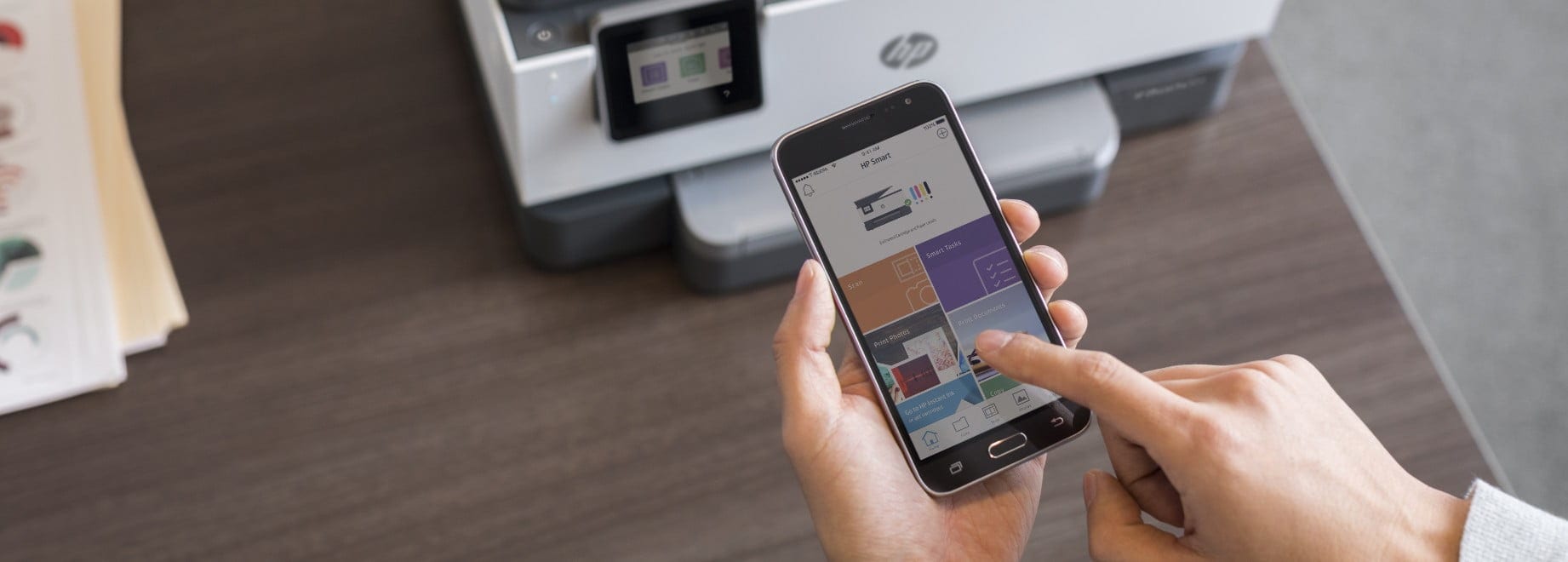 Hand tapping phone screen with HP Smart app, HP printer in background