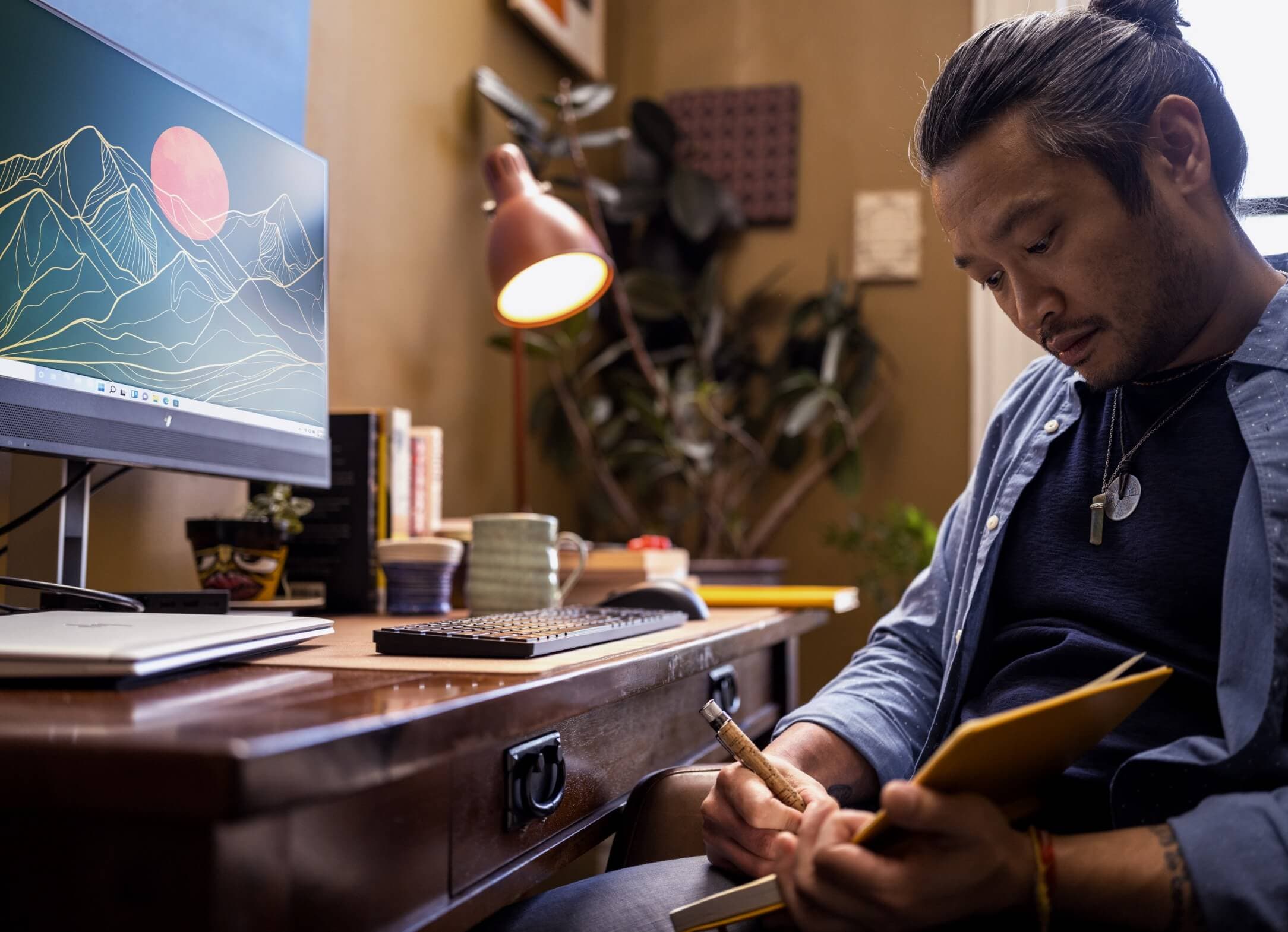 A man writting on a notebook aside an HP monitor, an HP wireless keyboard and mouse.