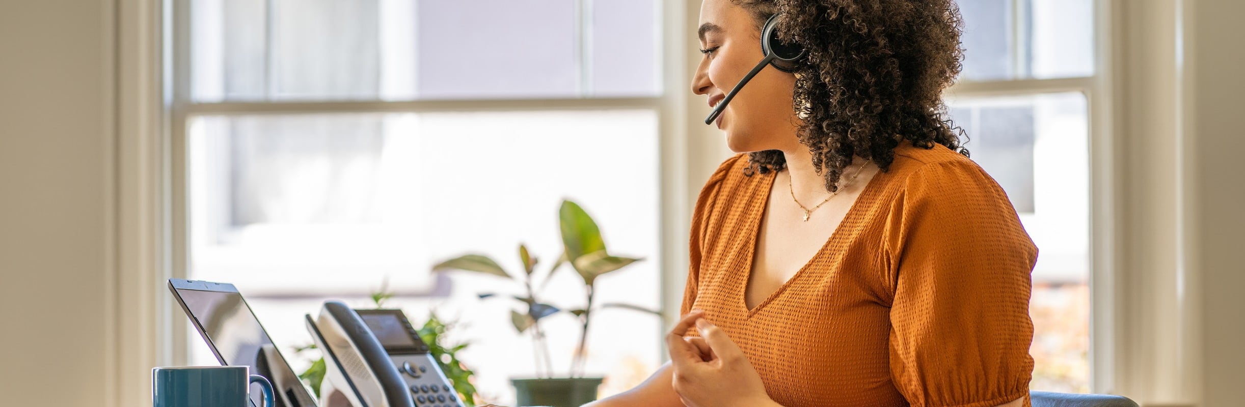 Woman seated at desk using headset, laptop and Poly Edge E300 desk phone​