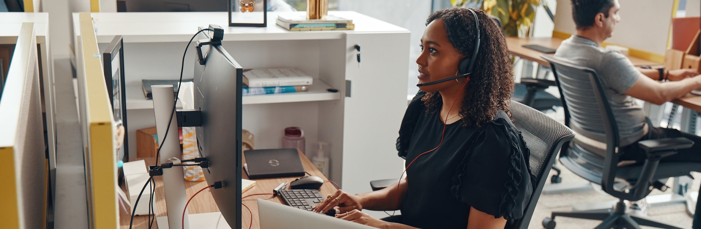 Woman seated at her office desk, wearing Poly Blackwire 3200 series headset while adjusting the webcam on the monitor during a video call