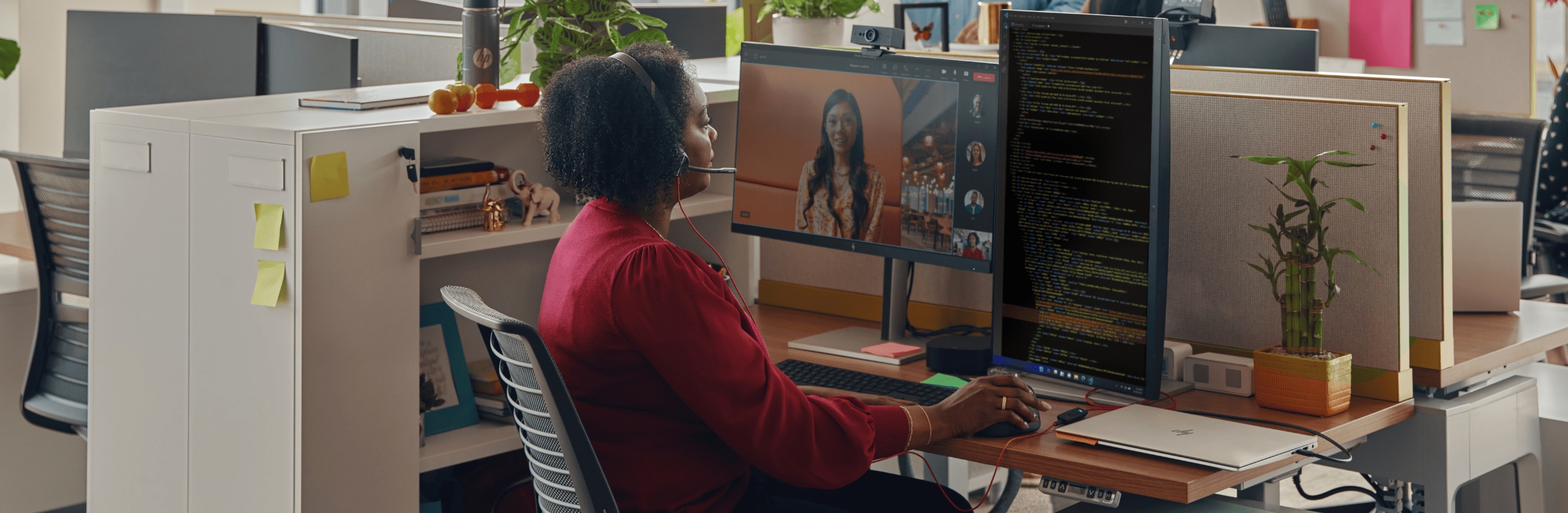 Woman seated at her office desk, wearing Poly Blackwire 3300 series corded headset while adjusting the webcam on her monitor during a video call