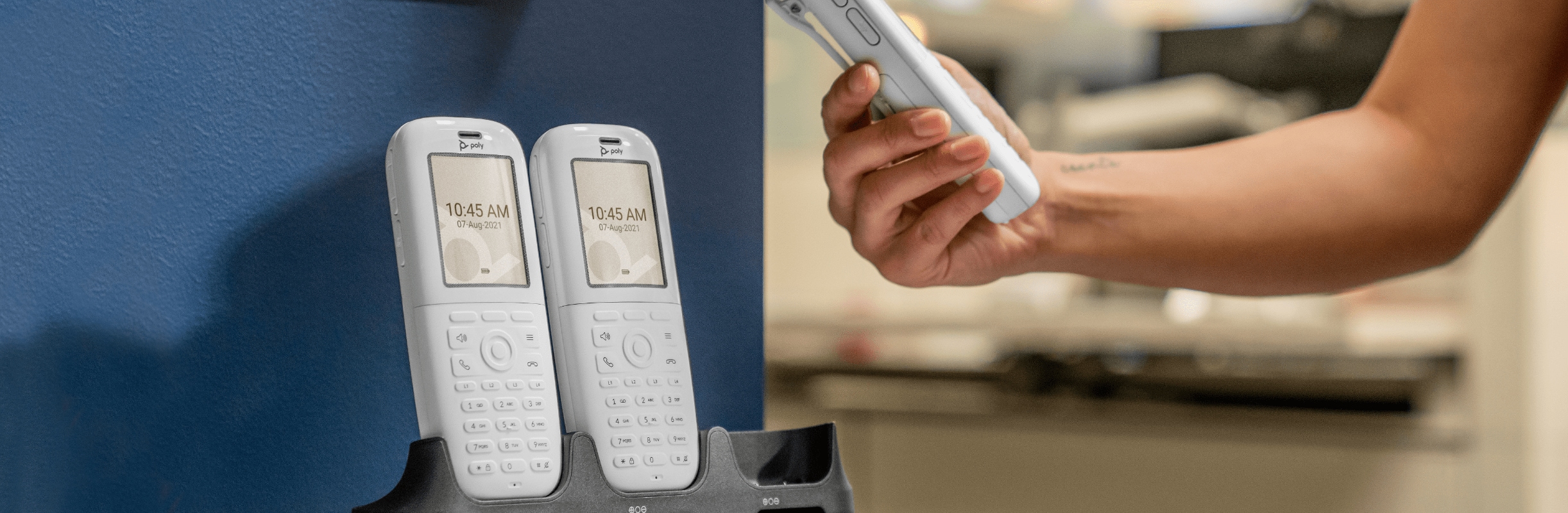 Person in office, holding a Poly Rove 40  wireless IP phone handset while two Poly Rove 40 handsets remain in base station beneath wall-mounted DECT repeater