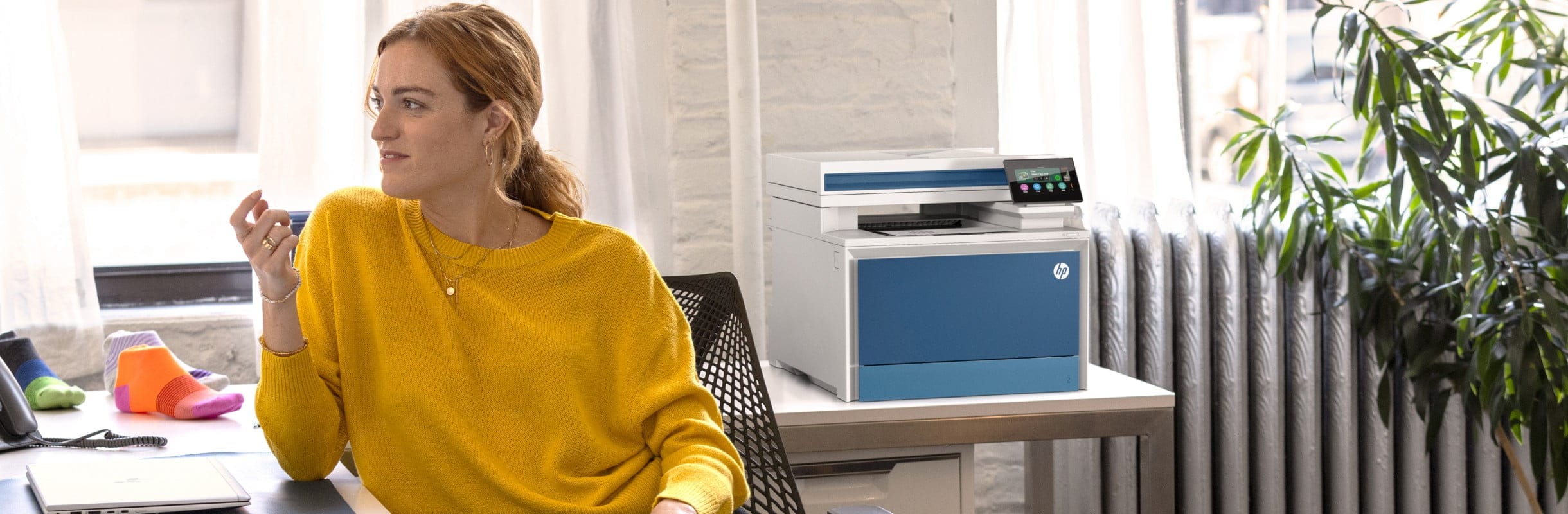 A woman is sitting in the office at a desk with an HP printer in the background