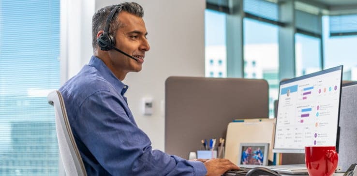 Man seated at office desk wearing Poly Savi 8210 headset while typing on keyboard and looking at Poly services screen on monitor