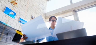 A person sorting through photography printouts in a modern, bright office with photos on the wall and large windows in the background.