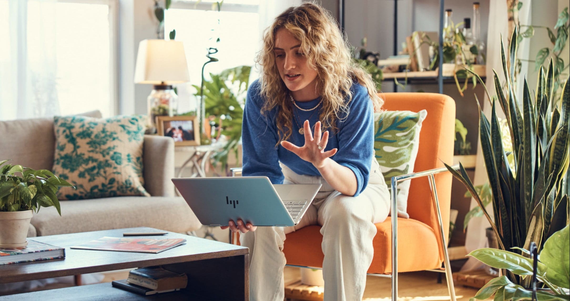 Woman in blue sweater sitting in an orange chair holding HP Spectre x360 laptop in one hand with other hand held up as if on a video call